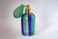 A vintage-style glass perfume atomizer bottle with vertical blue and green glass stripes, a gold metal collar, and a green mesh bulb pump, set against a plain light gray background.
