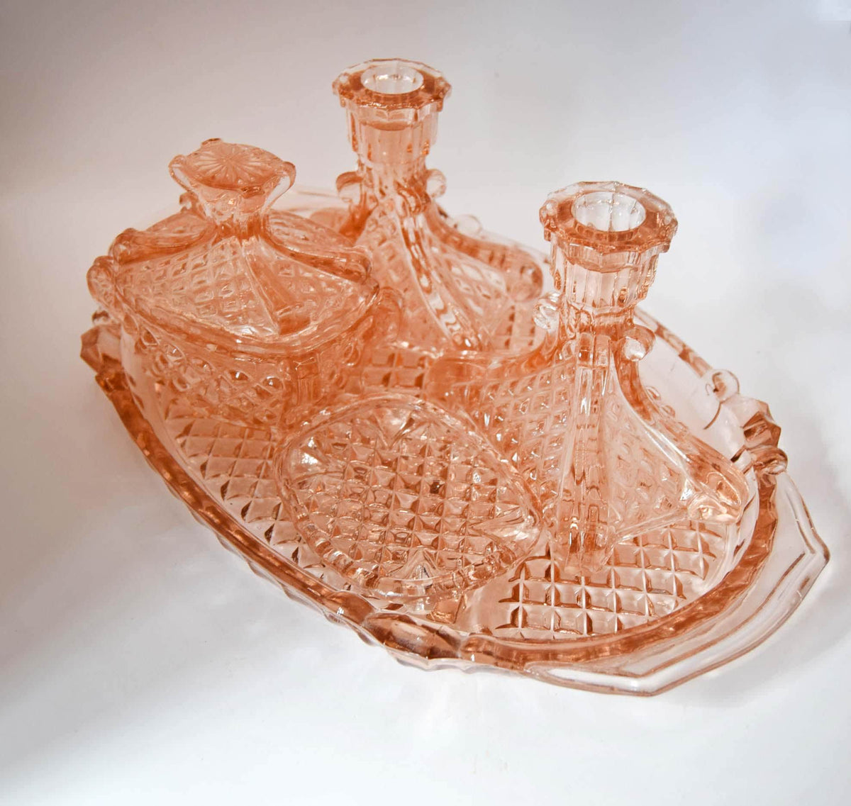 A vintage pink glass dish set featuring a rectangular tray with a diamond-cut pattern, a small lidded dish, and two candlestick holders, all in a translucent peach-pink color.