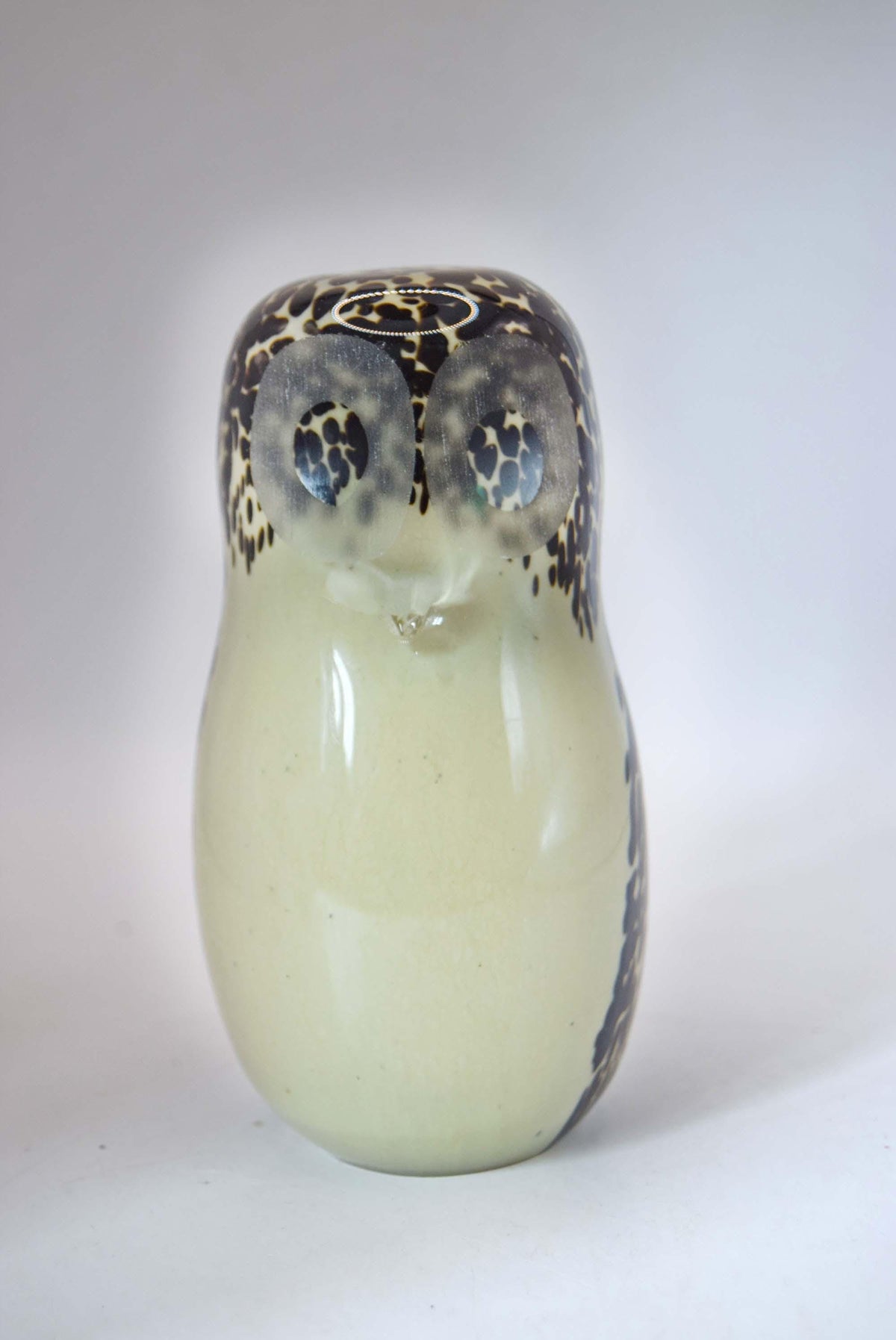 A ceramic owl figurine with a glossy cream-colored body and dark brown and black spotted details on its head and sides, set against a plain light gray background.