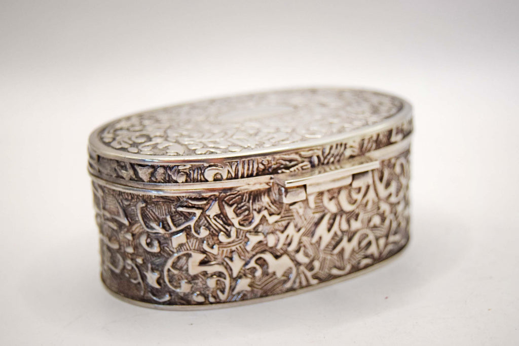 An ornate oval silver box with intricate floral engraving and a hinged clasp, photographed on a plain light background.