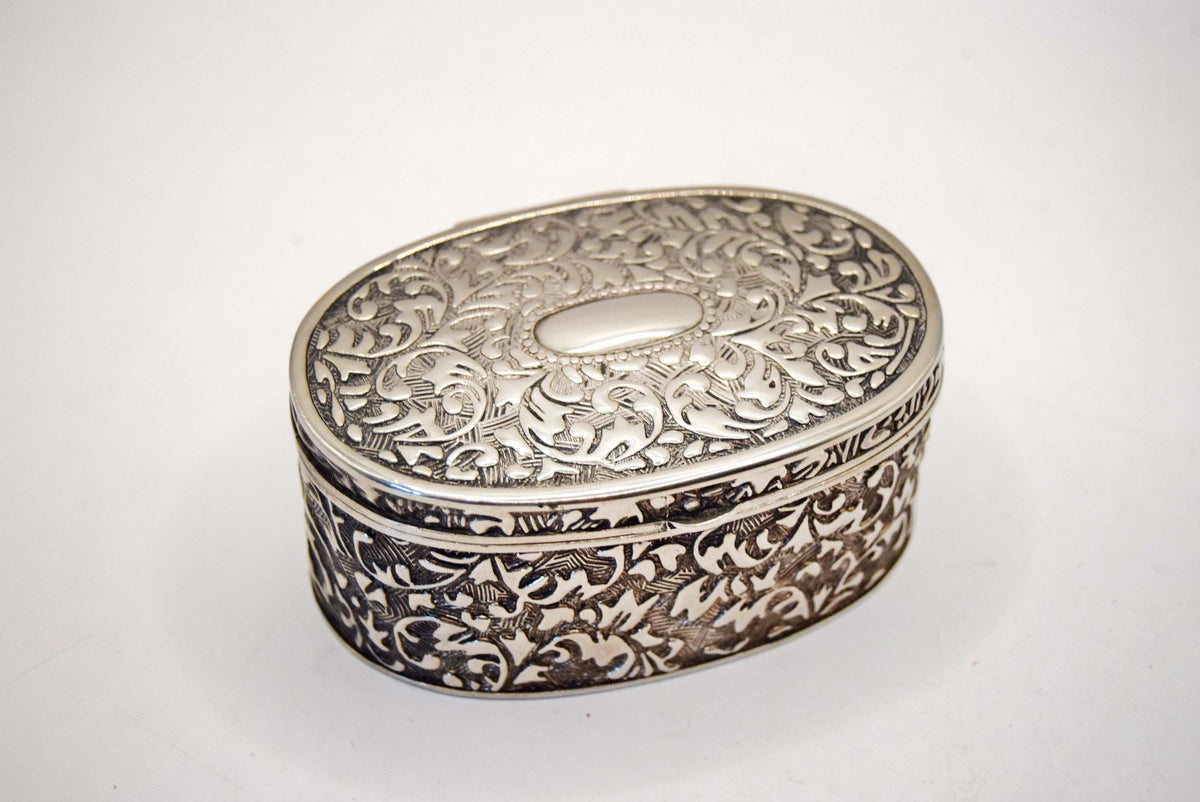 An ornate oval silver box with intricate floral engravings on the lid and sides, photographed on a plain white background.