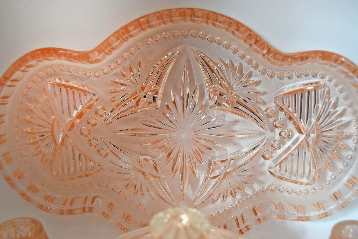 This 1930s–1940s Hollywood Regency Art Deco vanity set is crafted from heavy peach-pink depression glass featuring a geometric starburst and radial fan pattern.