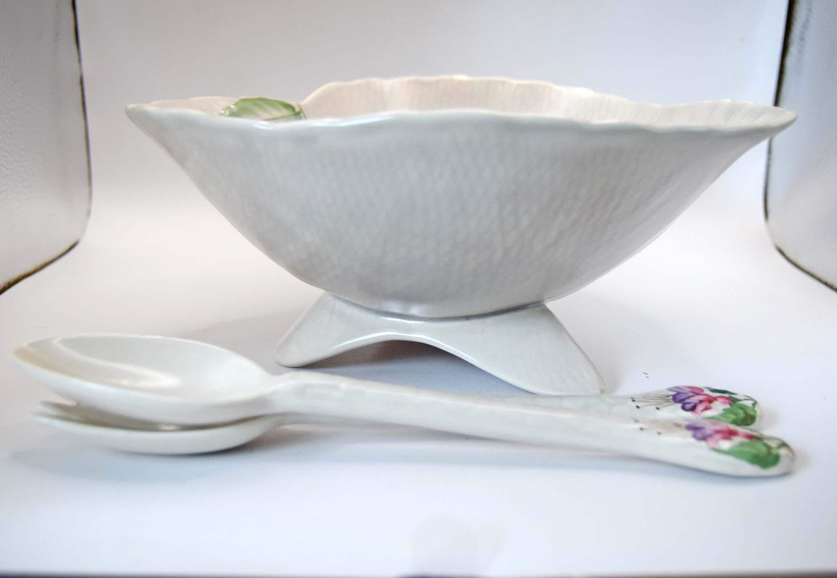 A white ceramic bowl with a textured surface and scalloped rim, shown on a matching pedestal stand with two decorative spoons featuring floral handles, set against a plain white background.