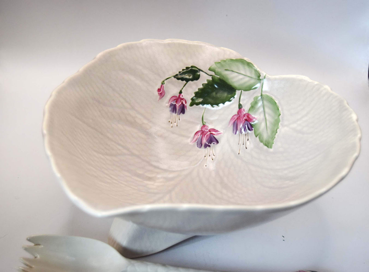 A white ceramic bowl with a leaf-like scalloped rim and embossed texture, decorated with painted pink-purple flowers and green leaves on the upper right side, shown on a white background with a white spoon partially visible at the bottom.