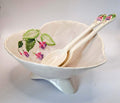 A white ceramic footed bowl with a scalloped rim, decorated with painted green leaves and pink flowers, accompanied by two matching ceramic spoons with floral handles, set against a plain light gray background.