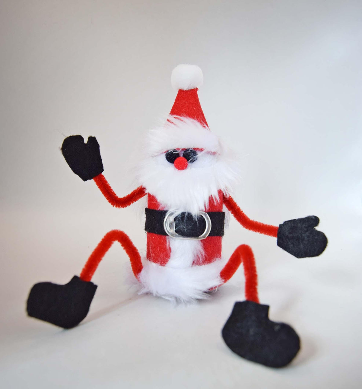 A handmade Santa Claus figure made from a red and white cylindrical container, decorated with a red felt hat, fluffy white faux fur beard, black felt gloves and boots, and a black belt with a silver buckle.