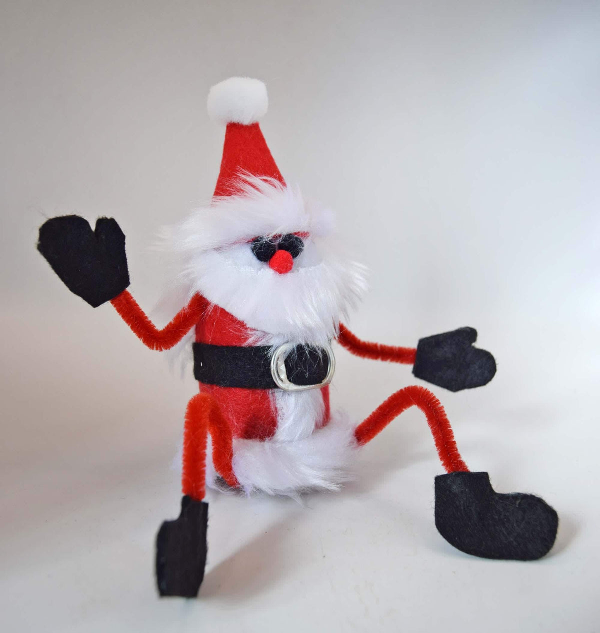 A handmade Christmas Santa Claus figure made from pipe cleaners, faux fur, and felt, posed sitting down against a plain light background.