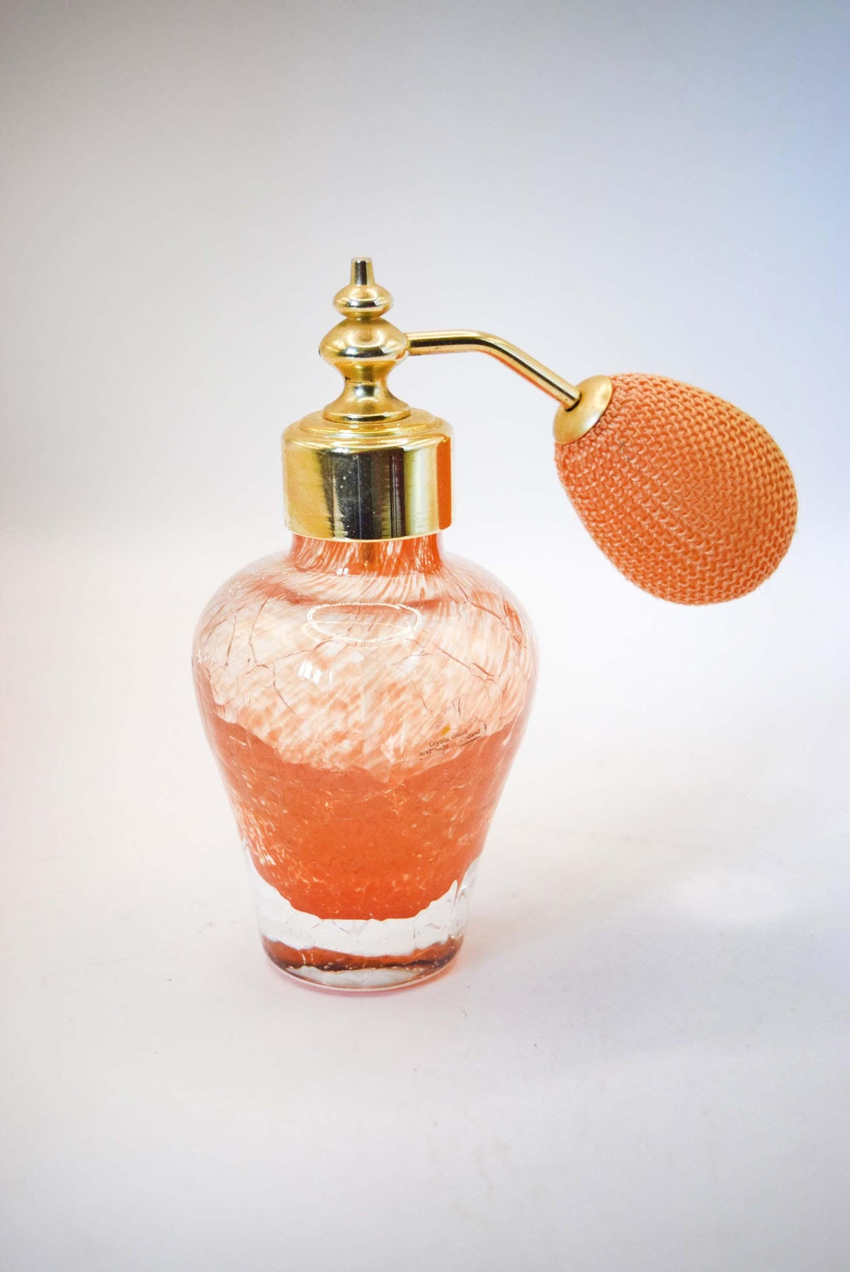 A vintage-style perfume atomizer bottle with a textured orange glass body, a shiny gold metal pump collar and nozzle, and an orange mesh bulb, shown on a plain light background.