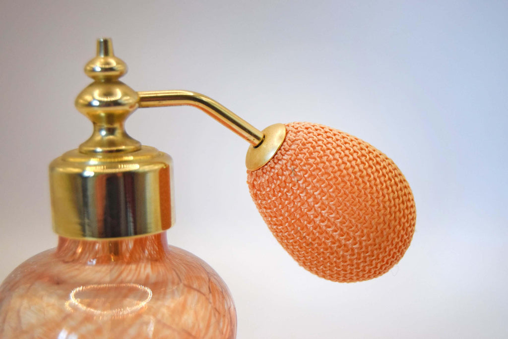 A close-up of a vintage-style perfume atomizer pump bottle with a gold-tone metal sprayer and a knitted peach-colored bulb, set against a soft light gray background.