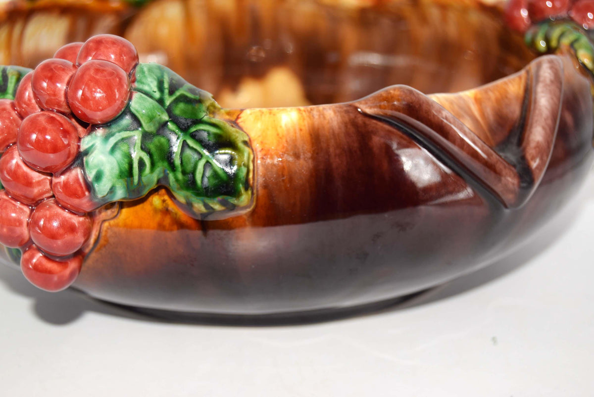 A close-up of a decorative ceramic bowl featuring a glossy brown and amber marbled glaze, with sculpted red grapes and green leaves on the left side, set against a plain white background.
