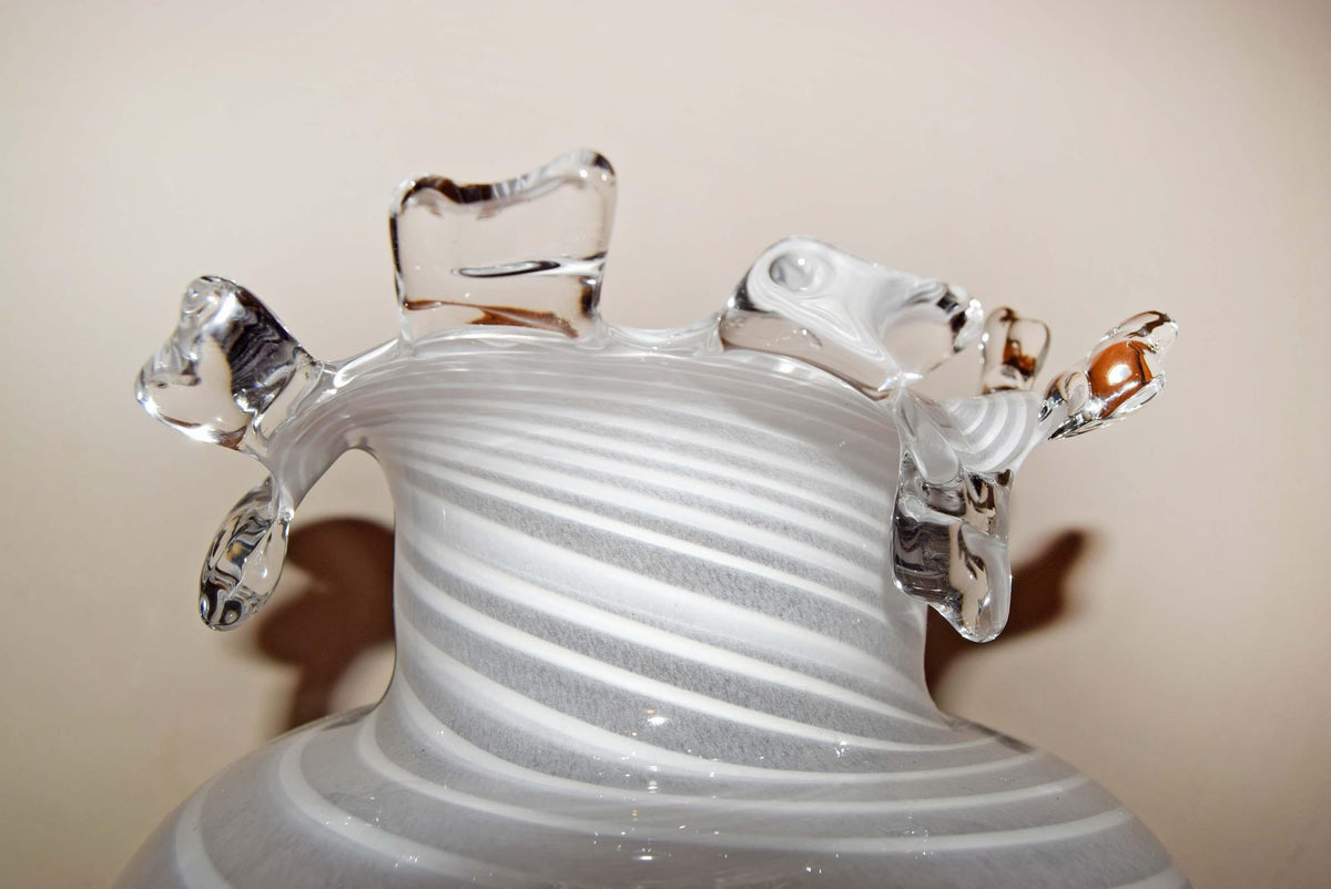 A close-up of a hand-blown glass vase with a flared rim, featuring horizontal swirled white and clear glass bands and three irregular transparent glass handles on a beige background.